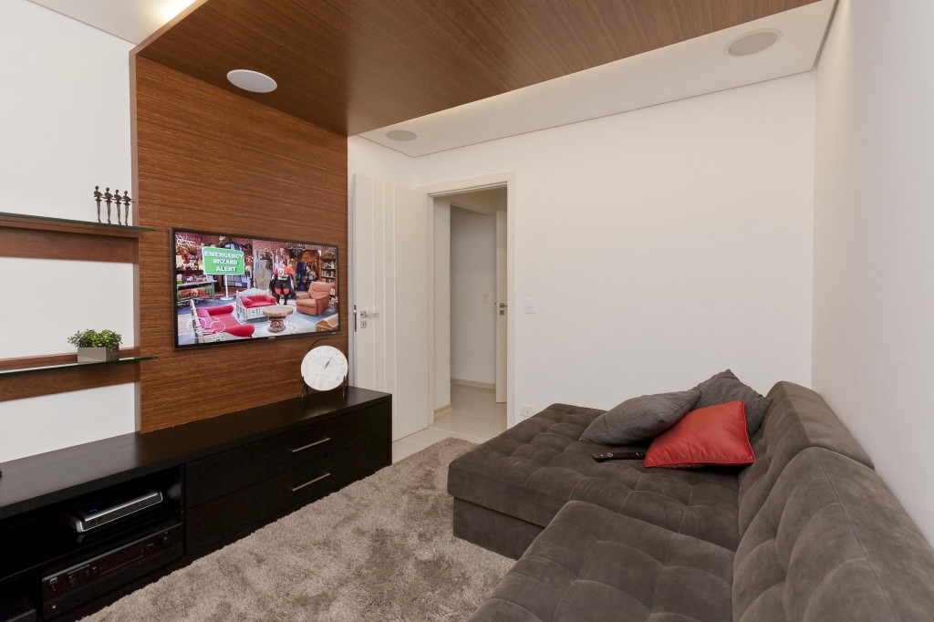 No home theater, o pórtico cumaru sustenta ao aparelho de TV e embute fita de led. O rack de MDF laqueado da Marcenaria Artesanal tem nichos abertos e gaveteiros para guardar os eletrônicos. O sofá com chaise (Breton) revestido de suede e o tapete (Santa Mônica) dão aconchego ao ambiente.