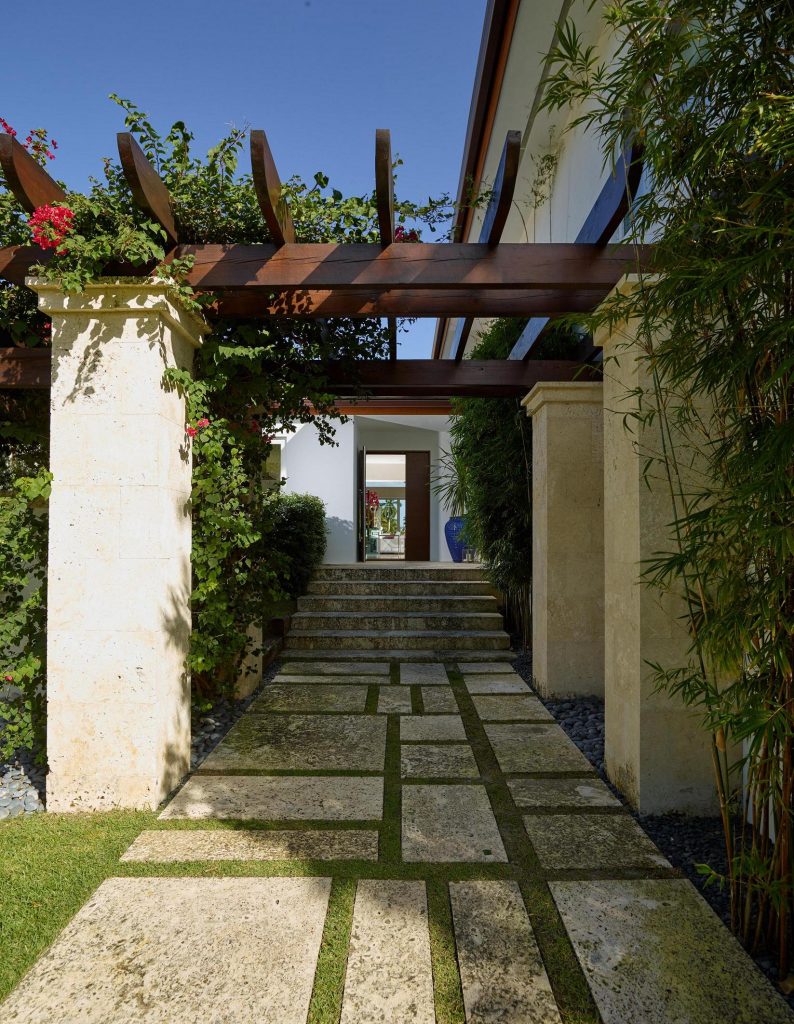 O paisagismo explora cada canto do terreno de 6.500 m2, como esse corredor lateral que leva à entrada da casa. Repare como o pergolado de madeira fornece sombra e frescor.